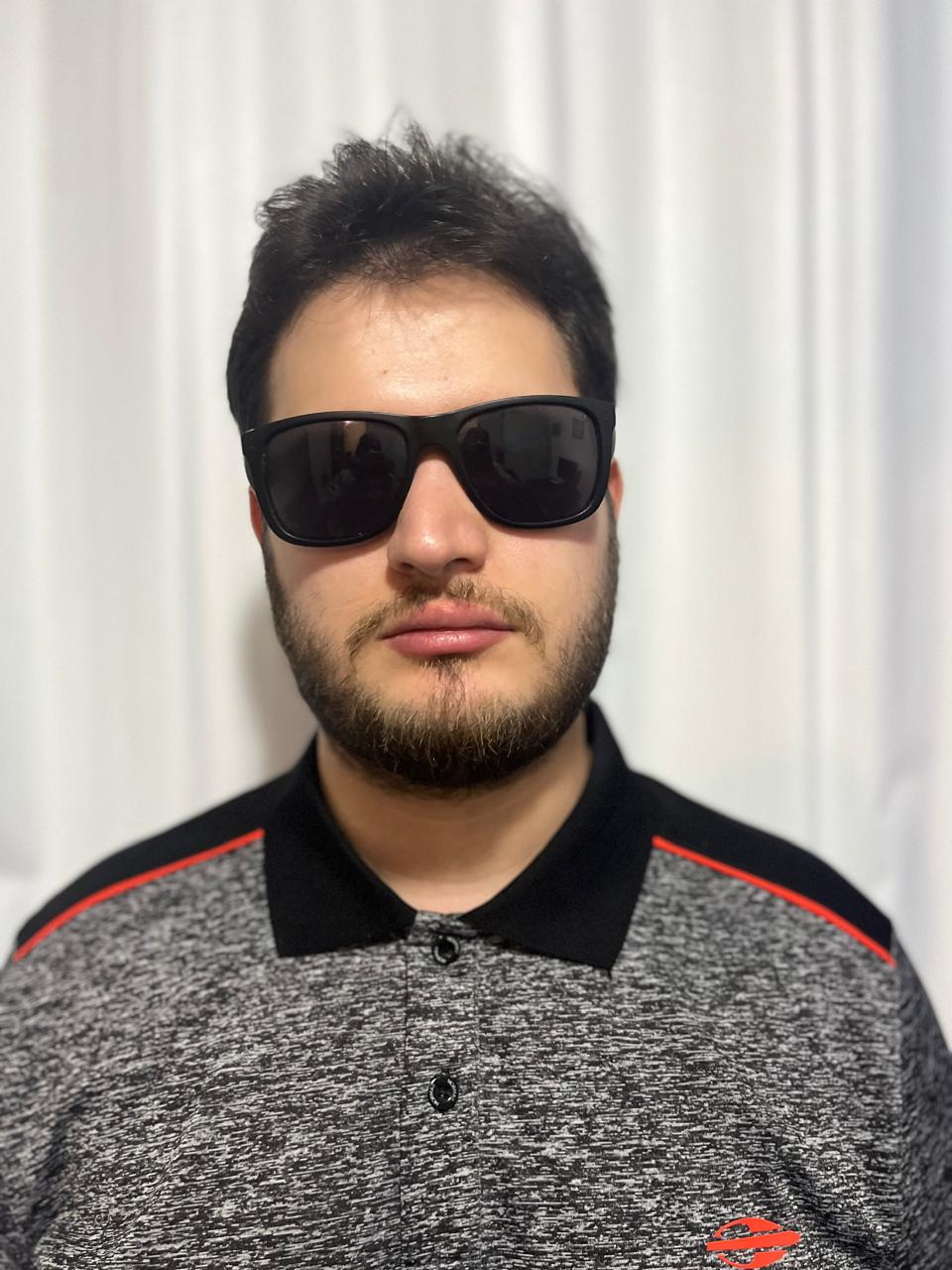 Imagem de uma pessoa com cabelo curto escuro e barba, usando óculos de sol e uma camisa polo cinza com detalhes em preto e laranja.